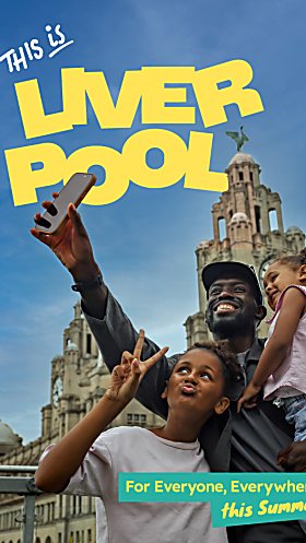 A family with an adult and two young children taking a selfie with the Royal Liver Building in the background. The text says 'This is Liverpool' in yellow text.