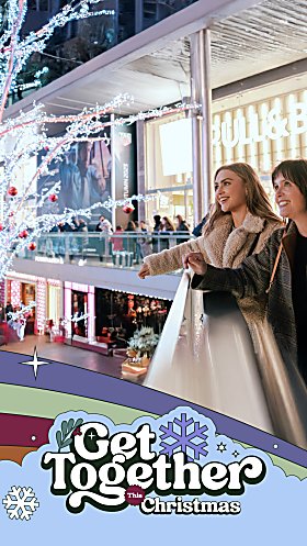 Two people stood next to a row of shops leaning over a railing looking at a large Christmas tree decoration.
