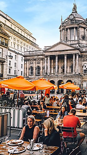 Liverpool Town Hall in the background of a shot of people eating a drinking at tables on a street. There are large orange umbrellas next to some tables.