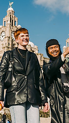 Two people infront of the Royal Liver Building similing. One is pointing at something off camera.