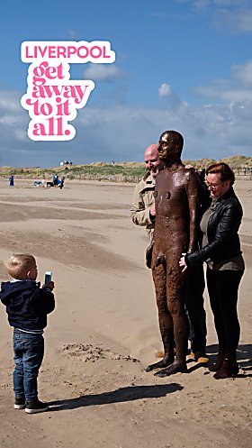 A young child is taking a picture of an adult male and female with a cast-iron, life-size statue on Crosby Beach.