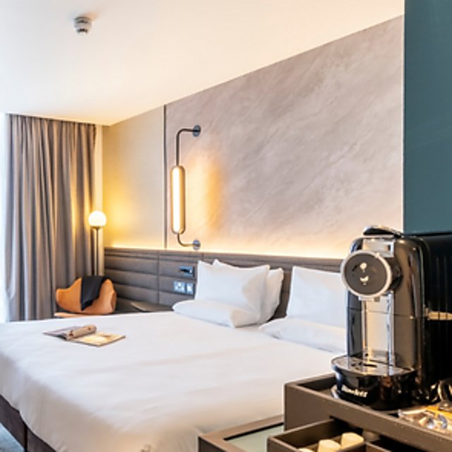 An extra large white bed in a spacious, airy room. The walls are a grey marble on one side and a deep green on the other. To the right is a special coffee machine and nearby a selection of coffee pods.