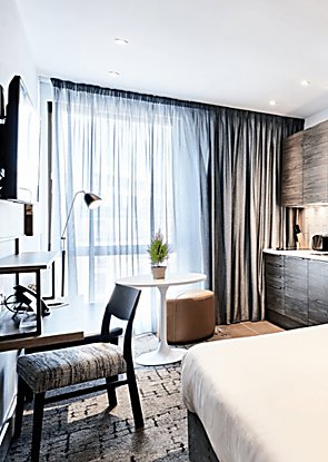 the view across a modern apartment. The room is light with white and grey details including a small kitchen area, large window and floor length curtains and a wall mounted television.