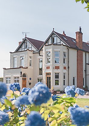 A large manor house painted white with huge windows and two pointed rooves. The photo is taken amongst blue flowers on a sunny day.
