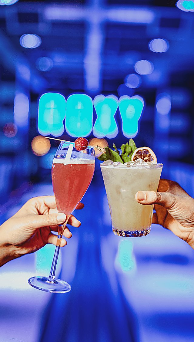 Two people 'cheers'ing two cocktails in front of a neon sign saying 'ROXY'.