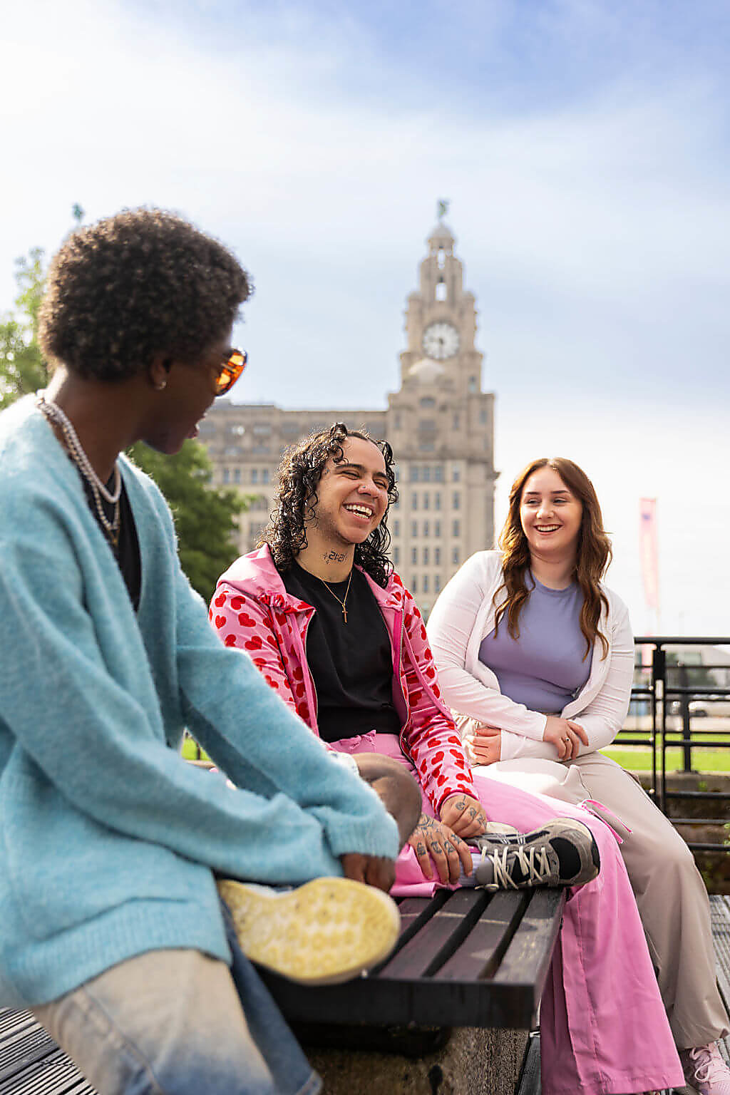 3 people sit on a bench on Liverpool Waterfront with the Royal Liver Building in the background