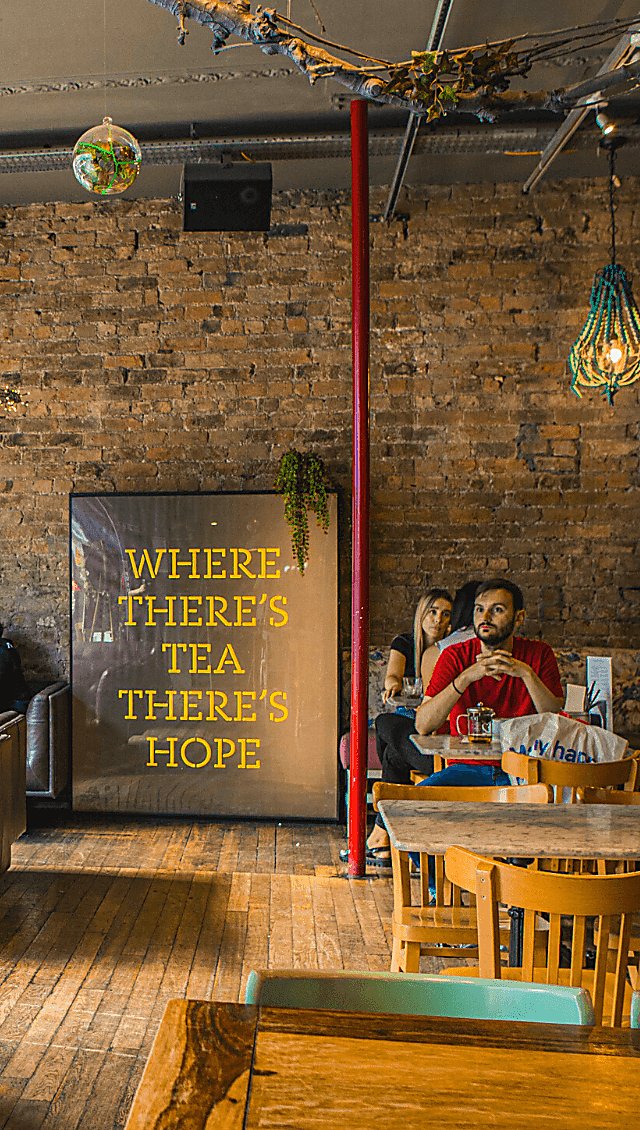 Inside the LEAF cafe. Against an exposed brick wall is a large print that reads 'where there's tea there's hope.' People are sitting at wooden tables and chairs in various chalky colours. There's also a disco ball hanging from the ceiling and a large red supporting column.