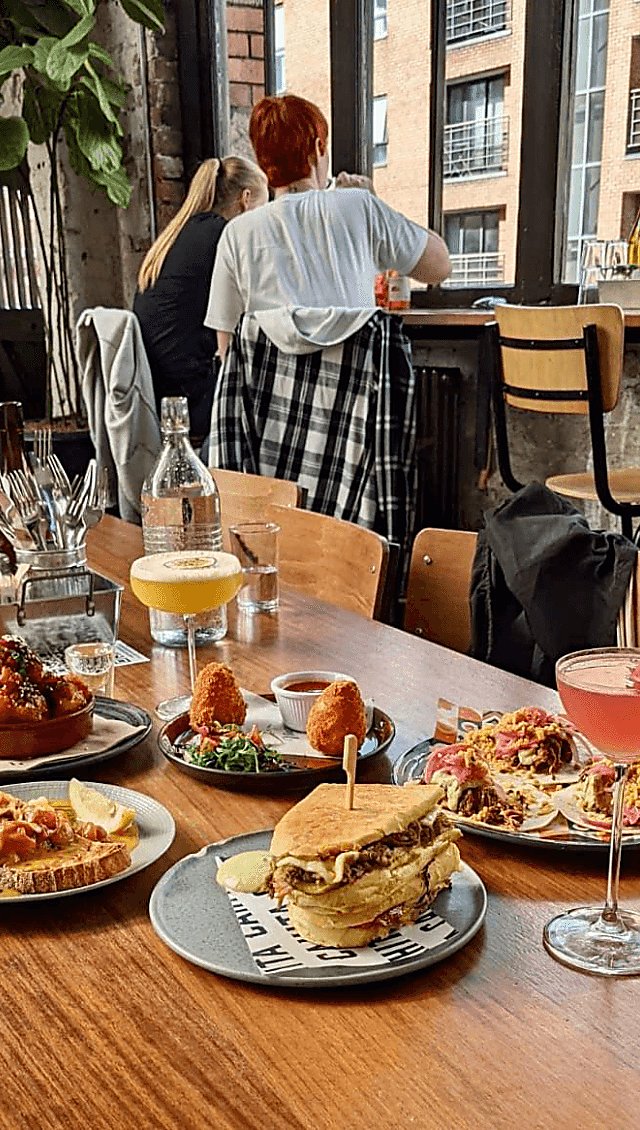 A selection of food on small plates. The food is served with two bright cocktails in martini style glasses. The room is light and airy and has bench seating.