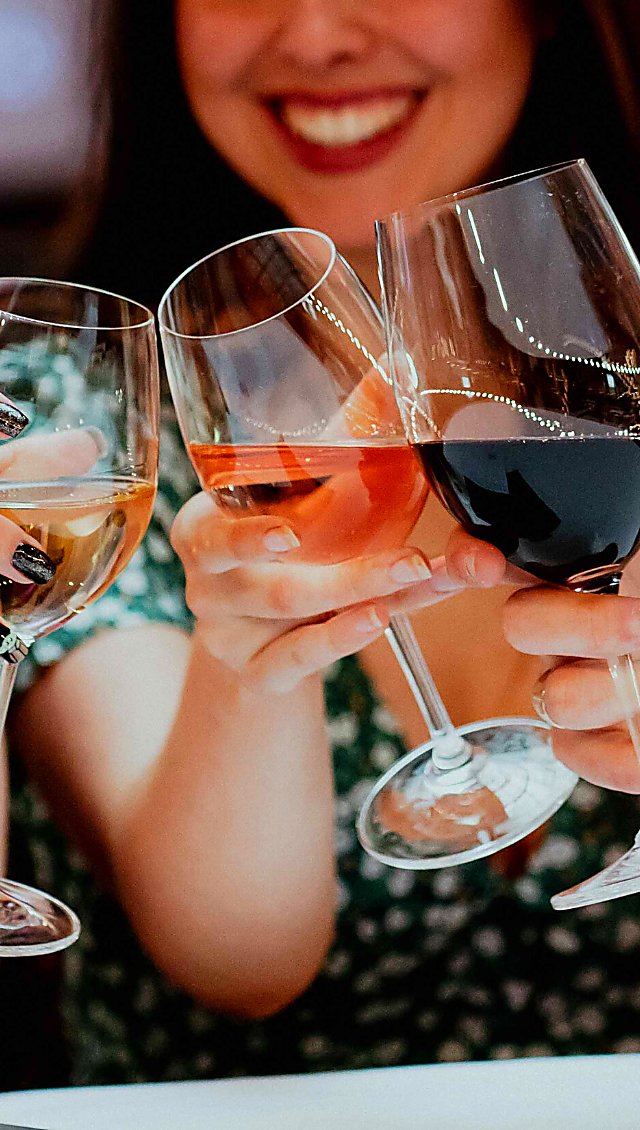 3 wine glasses filled with white, rose and red wine in a 'cheers' position over a table.