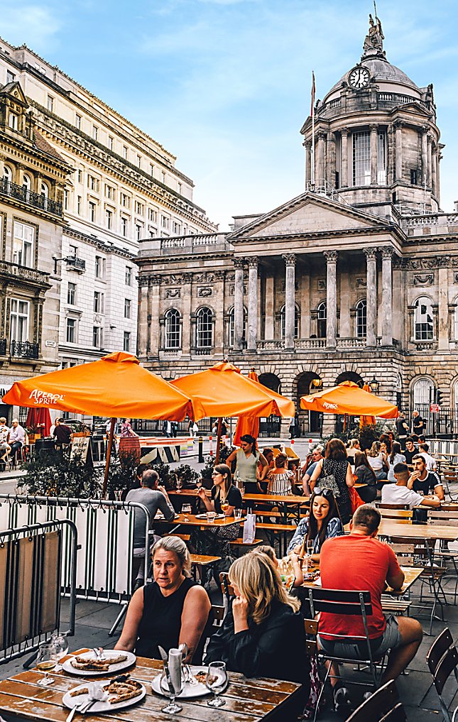 Liverpool Town Hall in the background of a shot of people eating a drinking at tables on a street. There are large orange umbrellas next to some tables.
