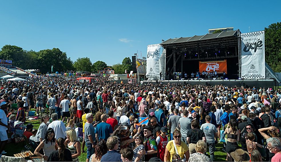 Wide outdoor festival scene with a large crowd standing on grass in front of a main stage. The stage features lighting rigs, banners, and a large central screen, with trees and fairground rides visible in the background under a clear blue sky.
