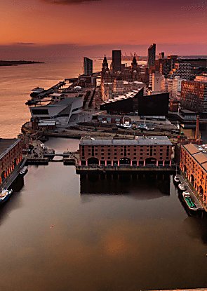 A drone shot over Liverpool looking across Royal Albert Dock warehouse buildings and the Pier Head with orange sky