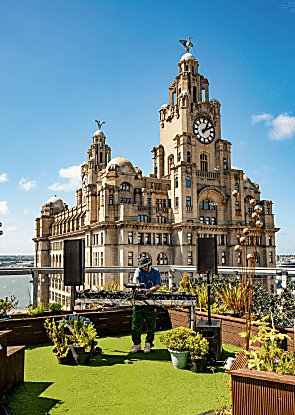 A person standing on a rooftop with the Royal Liver building behind them, they're playing music and the sky is blue.