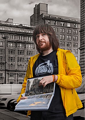 A man standing with a booklet showing a film location. He is wearing a bright yellow coat and pointing to the photo.