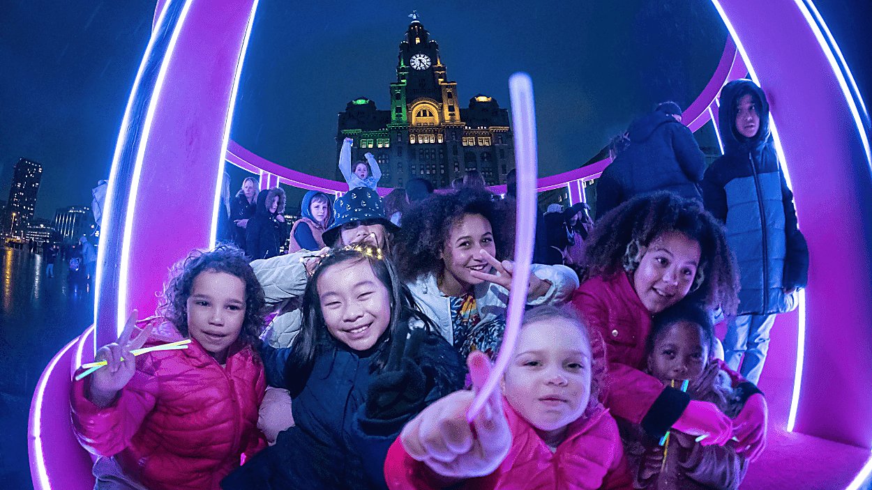 A group of children inside one of the light installations with the royal liver building in the background.