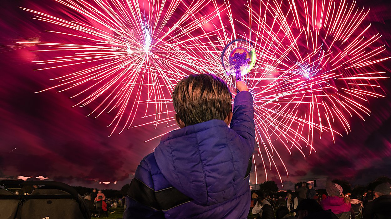 A boy holding up a toy light with fireworks being let off in the distance.