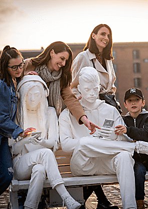 Two adults and two children engage with a statue of people painted white, sitting on a bench looking at 'smart phones.'