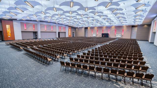 Large indoor ballroom with several rows of chairs placed in a theatre style arrangement facing a small stage and tv screens on each side of the stage.