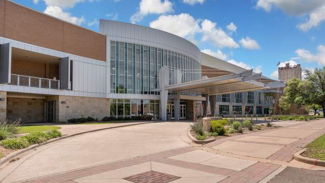 Waco Convention Center exterior with modern glass facade, curved entrance canopy, landscaped walkway, blue sky, clouds, Texas day.