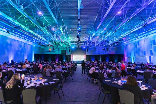 Colorful Uplighting and Attendees at Tables in Front of a Stage for a Conference Presentation
