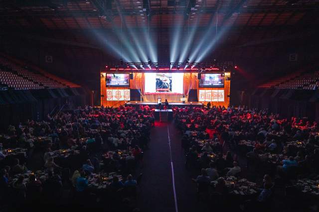Stage and projector screens on the back of the arena floor with round guest tables for banquet attendees on the remaining floor space