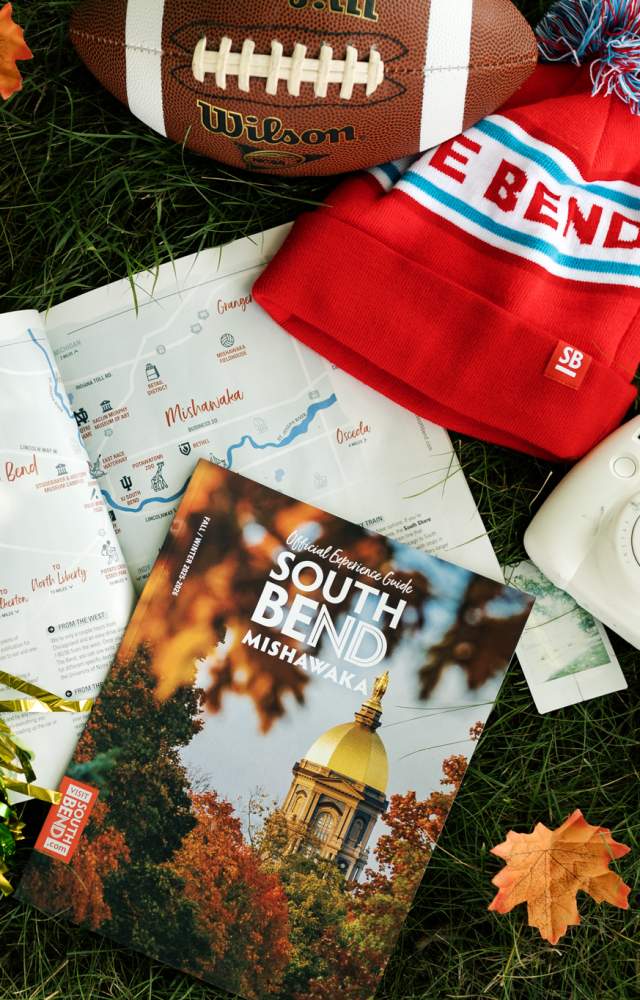 A copy of the 2025-2026 Fall/Winter Experience Guide featuring the University of Notre Dame's golden dome amongst fall foliage lies in the grass on campus, surrounded by a green and gold pom-pom, a winter hat that says "The Bend", a football, and a Polaroid camera. Fall leaves blow gently across the scene with the breeze.