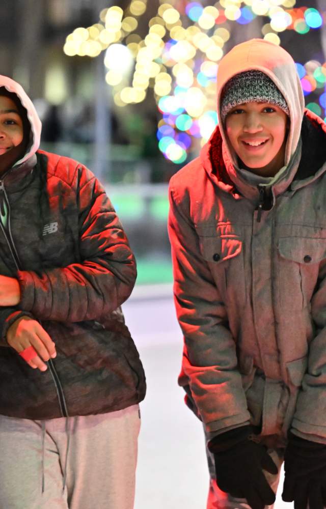 Two young boys smile while taking a break from ice skating with holiday lights in the background