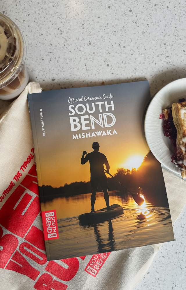 An issue of the 2025 Spring/Summer Experience Guide with a sunset image of a paddle boarder on a lake at Potato Creek state park lays on a counter of a coffee shop with an iced coffee, a pastry and a canvas tote bag.