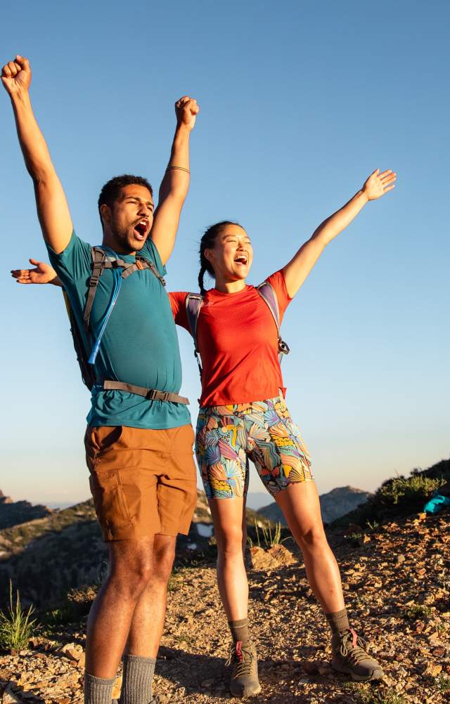 Man and Woman wearing hiking gear with their arms up and looking excited to reach the top of a mountain