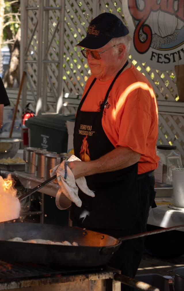 Two pyro chefs at the Gilroy Garlic Festival 2025 expertly cook over open flames, sending a dramatic burst of fire into the air from a giant skillet. One chef wears an orange shirt and apron that reads 'Heard You Missed Us!' as he stirs sizzling garlic shrimp.