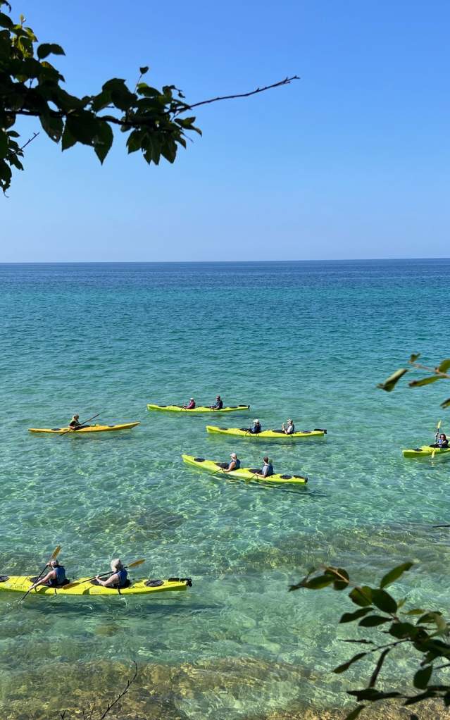 Kayakers paddle bright yellow kayaks in the clear turquoise water of Lake Superior, framed by leafy branches, under a cloudless blue sky, conveying a sense of calm.