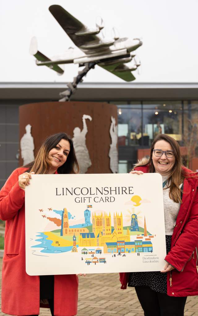 Two people holding a large Lincolnshire Gift Card in front of the International Bomber Command Centre