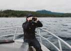 A man sits on a fishing boat with Nelson Island in the background, looking through binoculars.