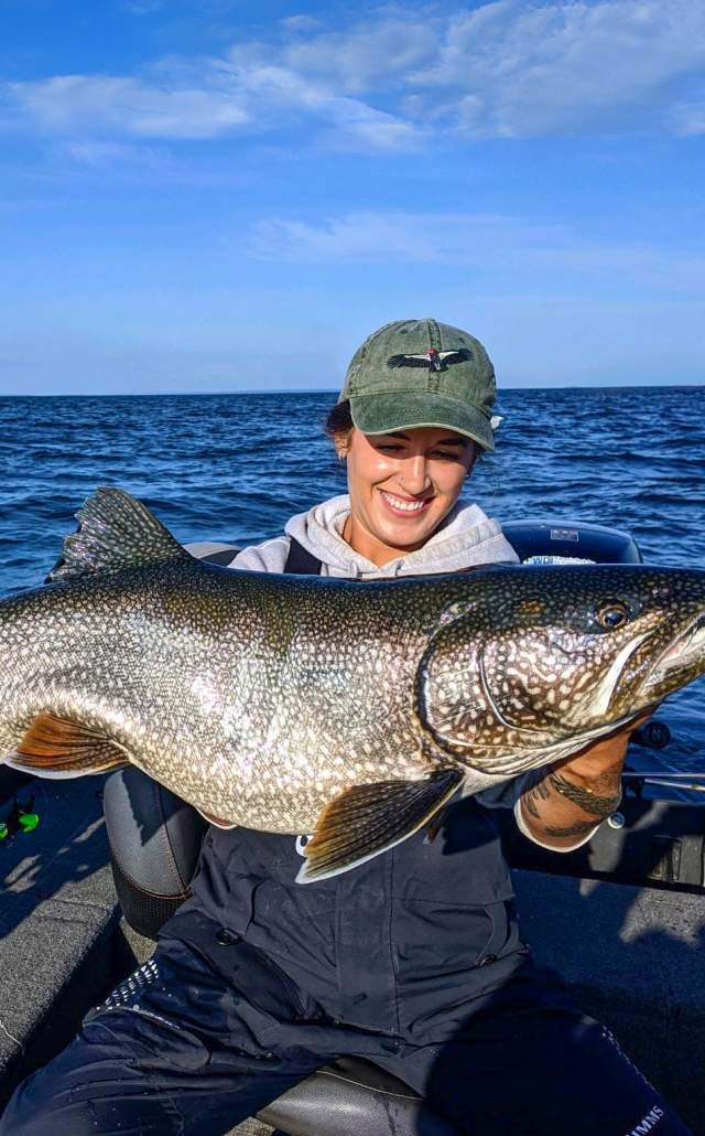 A person in a boat happily holds a large fish against a backdrop of the vast waters of Lake Superior under a clear blue sky, conveying excitement and achievement.