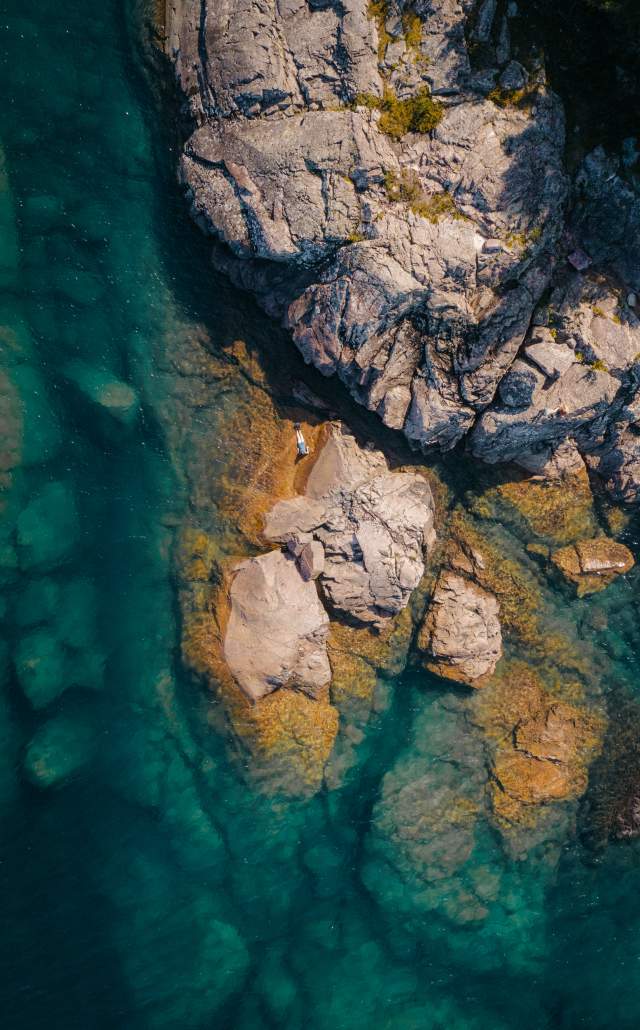 Aerial view of rocky cliffs meeting clear turquoise water along Lake Superior's shoreline, with sunlight highlighting the rocks beneath. Sparse greenery on the cliff adds a natural touch.