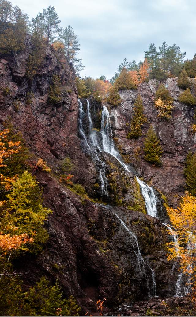 Cascading waterfall flows over rocky cliffs, framed by vivid red, orange, and golden autumn foliage beneath an overcast sky.
