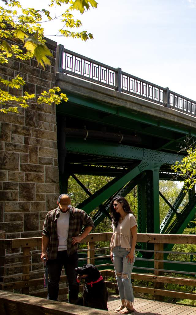 A man and woman stand with a black dog on a sunny wooden deck next to a green metal bridge and leafy trees, conveying a peaceful outdoor setting.