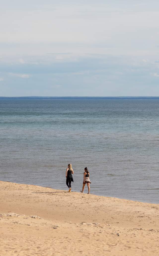 Two people walk along Lake Michigan on a sandy beach under a cloudy sky, conveying a peaceful, serene atmosphere. Distant land is visible on the horizon.