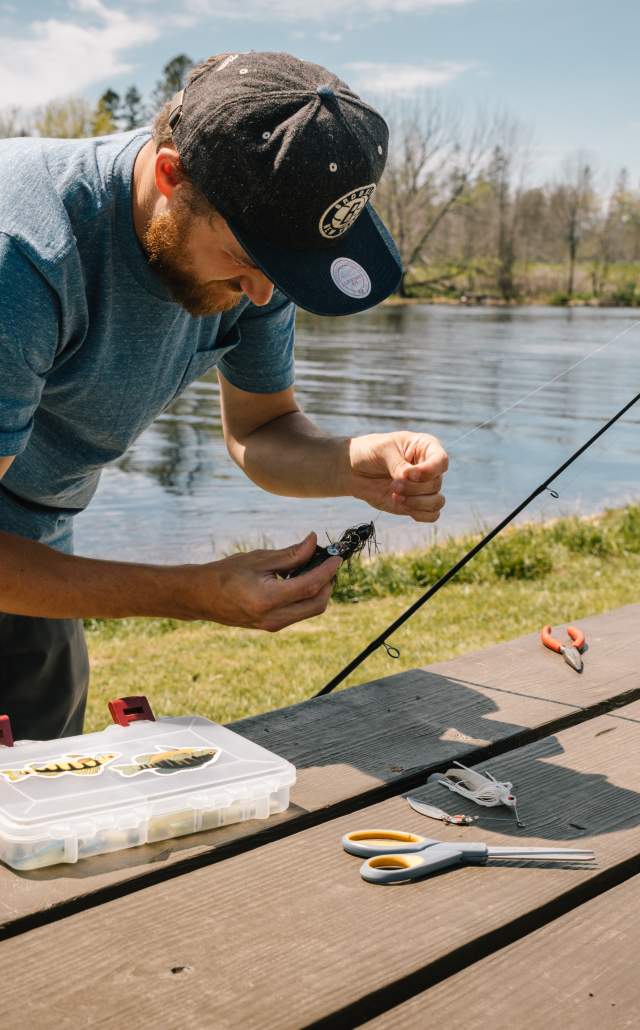 Man in a hat prepares fishing gear at a wooden picnic table by a river. Tackle box, scissors, and pliers are on the table. Sunny day with green trees.