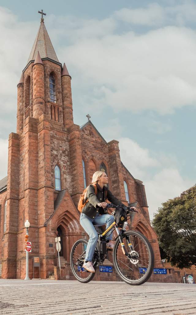 A person rides a bicycle past a large, historic brick church with tall spires and Gothic architecture. The sky is partly cloudy, creating a serene atmosphere.