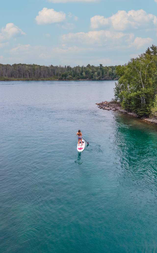 A person paddleboarding the clear turquoise water of the St. Marys River near a lush green shoreline and under a partly cloudy sky. The scene conveys tranquility and adventure.