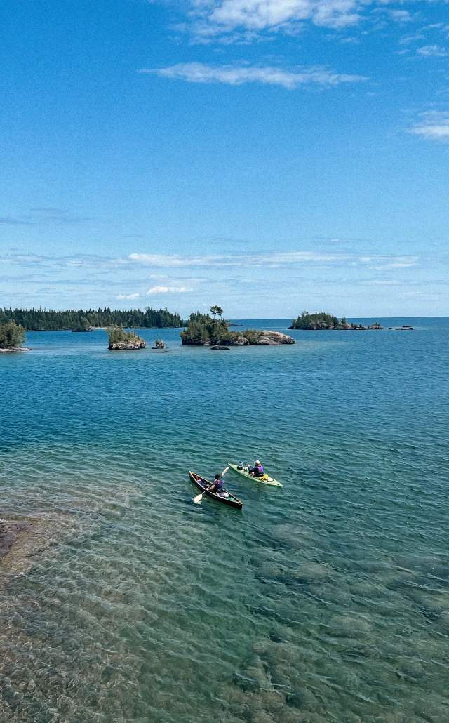Two people in kayaks paddle on clear blue water of Lake Superior near the rocky shoreline of Isle Royale National Park. Small, tree-covered islands are visible under a bright blue sky. Serene atmosphere.