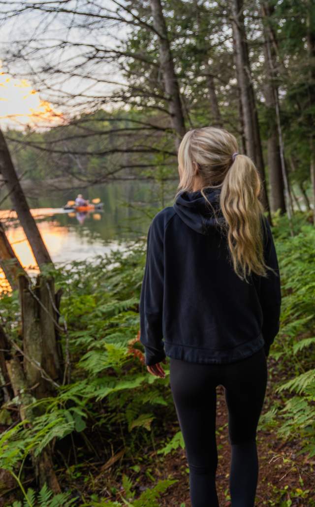 A woman with long blonde hair in a ponytail stands facing a serene lake in the Sylvania Wilderness at sunset, surrounded by lush green ferns and tall trees.
