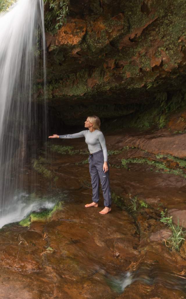 A young woman stands barefoot on a reddish-brown rock ledge, reaching toward the powerful, white curtain of a waterfall that flows over a mossy, rocky cliff face.