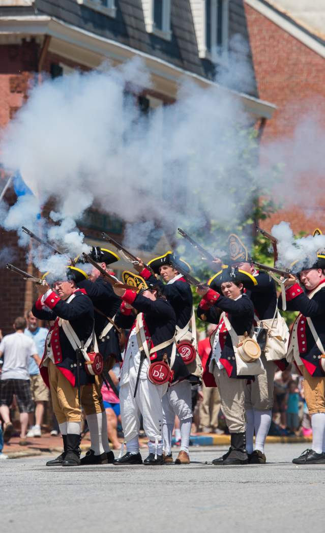 A group of historical reenactors in colonial military uniforms fires muskets, creating clouds of smoke during a lively outdoor event.