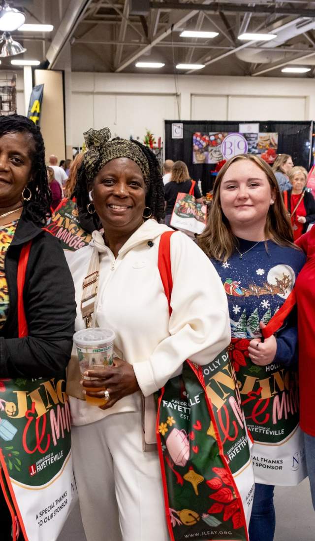 A group of smiling shoppers pose with festive Holly Day Fair tote bags while exploring vendor booths filled with holiday gifts and apparel at the Crown Expo Center in Fayetteville, NC.