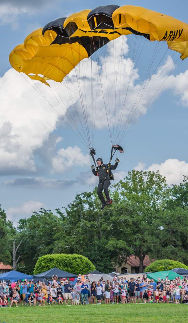 Paratrooper descending under a yellow parachute onto a parade field at Fort Bragg, with spectators watching from the sidelines.