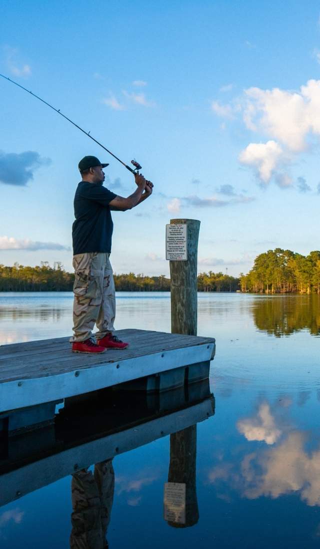 Person fishing from a wooden dock on a calm lake in the early evening, with trees and clouds reflecting on the water at Hope Mills Lake Fishing.