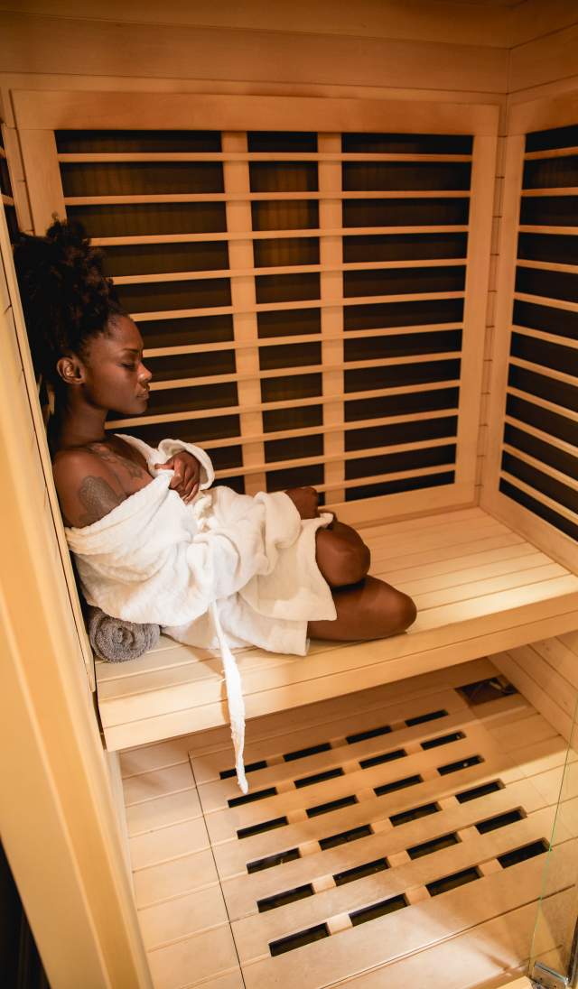 A black woman sits peacefully in a cedar sauna, wrapped in a white robe with eyes closed, enjoying a calming self-care moment in Fayetteville,
