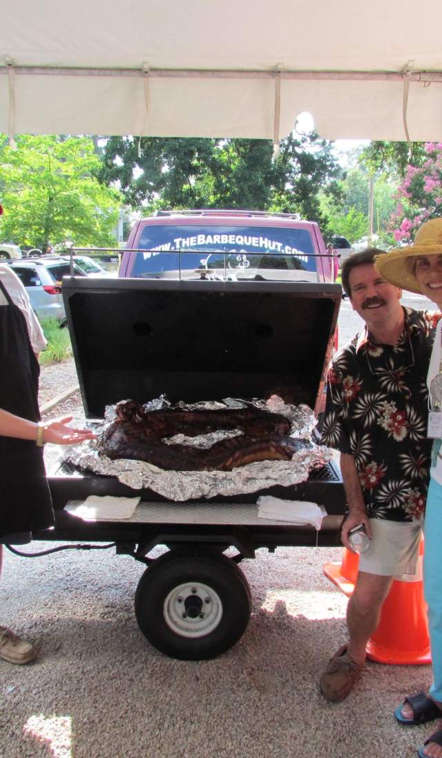 Group of adults smiling and gathered under a tent around a barbecue grill with smoked meat during an outdoor reunion-style event, creating a casual and social atmosphere.